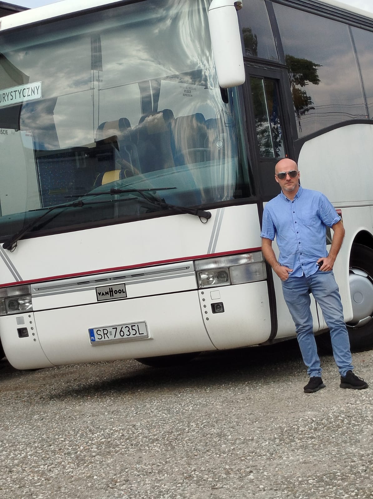 Biały autokar Van Hool z polską rejestracją SR-7635L, na tle szarego żwiru, z mężczyzną w niebieskiej koszuli i okularach przeciwsłonecznych, opierającym się o pojazd.