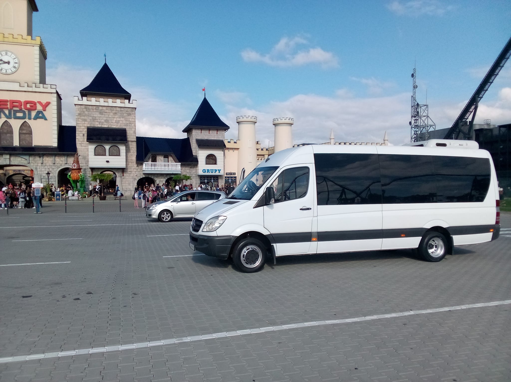 Biały bus na parkingu przed parkiem rozrywki EnergyLandia. Pojazd z przyciemnianymi szybami, widoczny z boku, na pierwszym planie. W tle budynki parku i spacerujący ludzie.