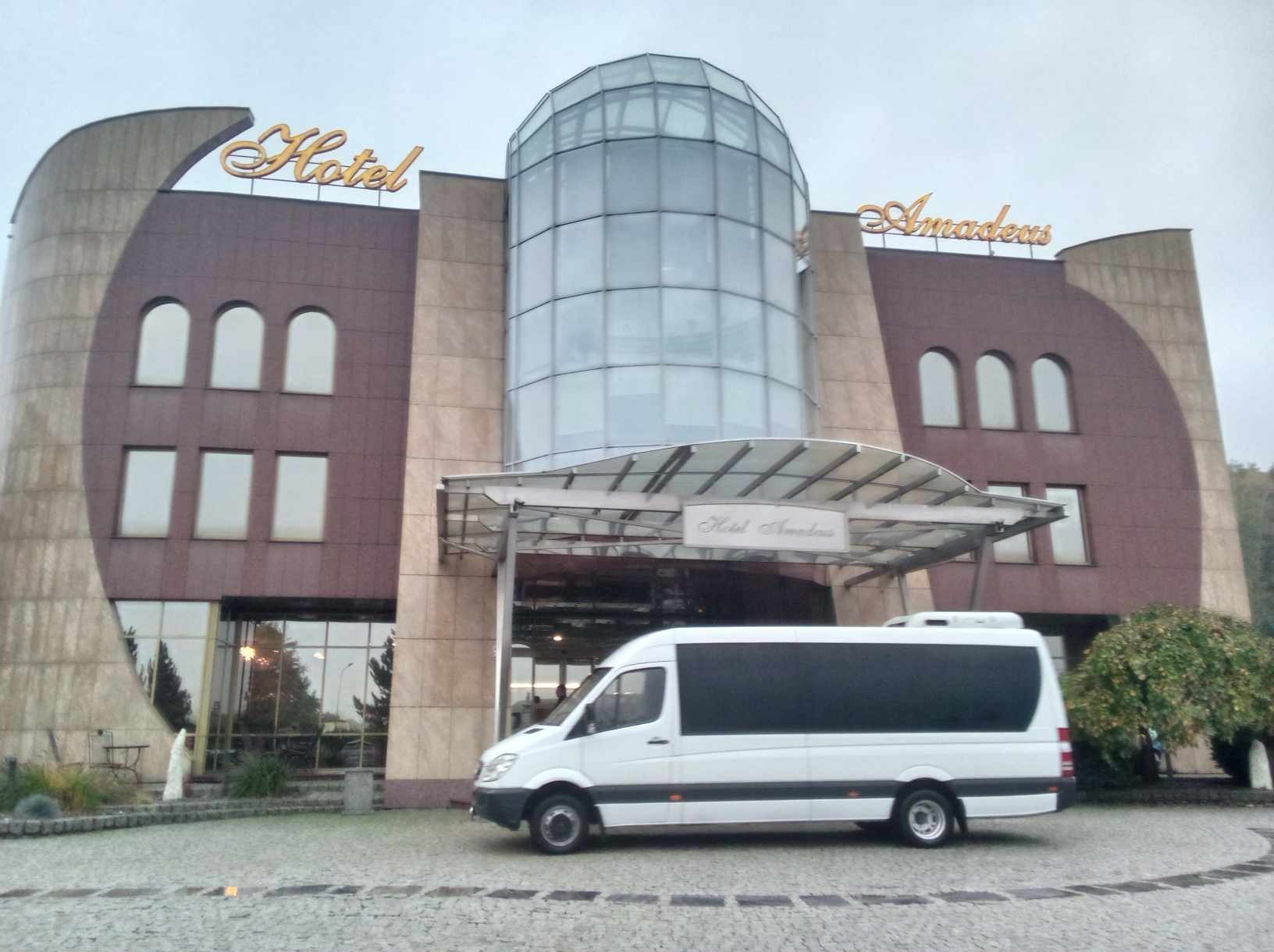 Biały bus z przyciemnianymi szybami zaparkowany przed hotelem Amadeus z przeszkloną kopułą. Transport gości hotelowych.