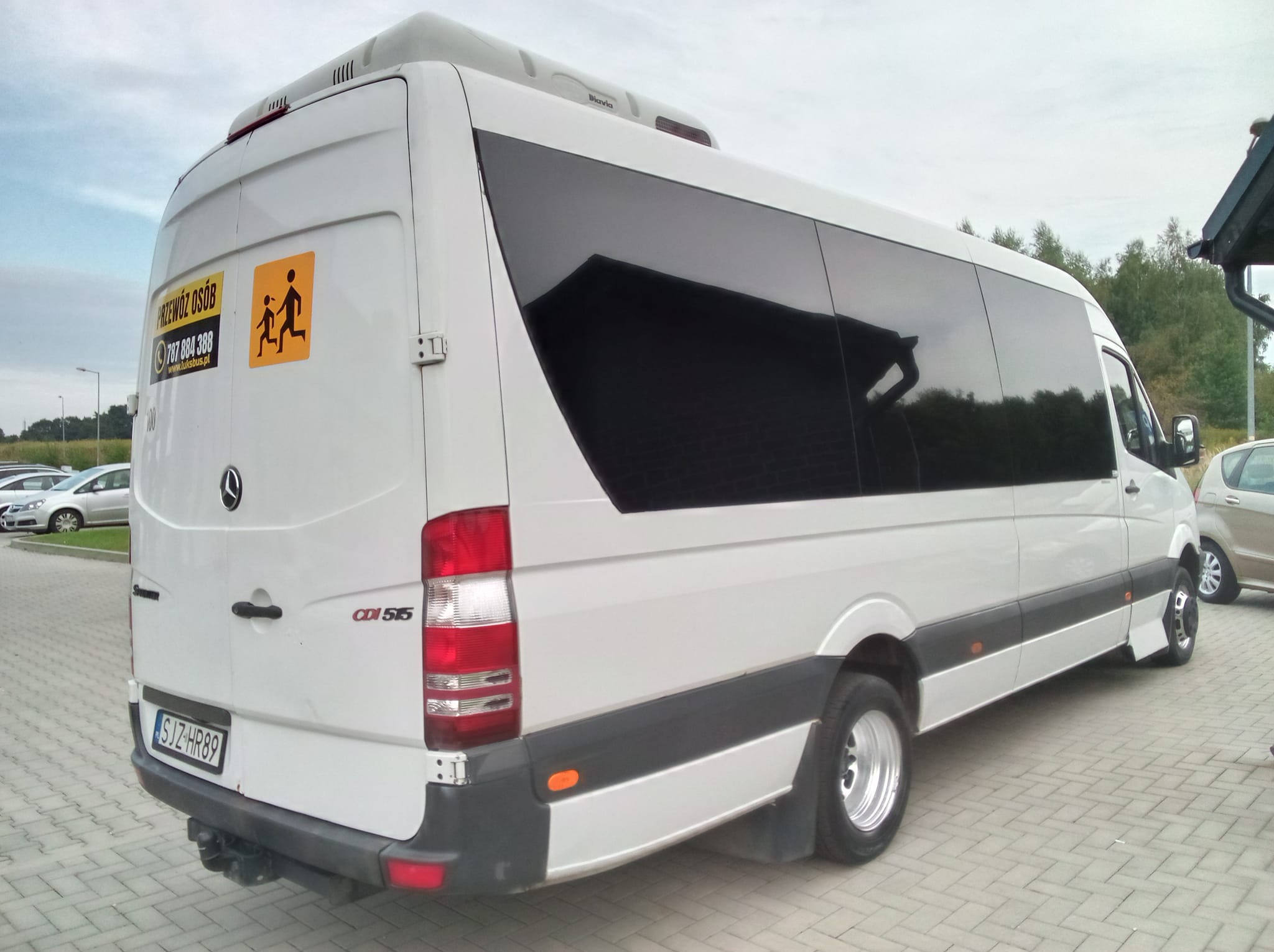 Biały bus Mercedes Sprinter CDI 515 z przyciemnianymi szybami i naklejką z symbolem dzieci, zaparkowany na kostce brukowej. Widok z tyłu i boku.