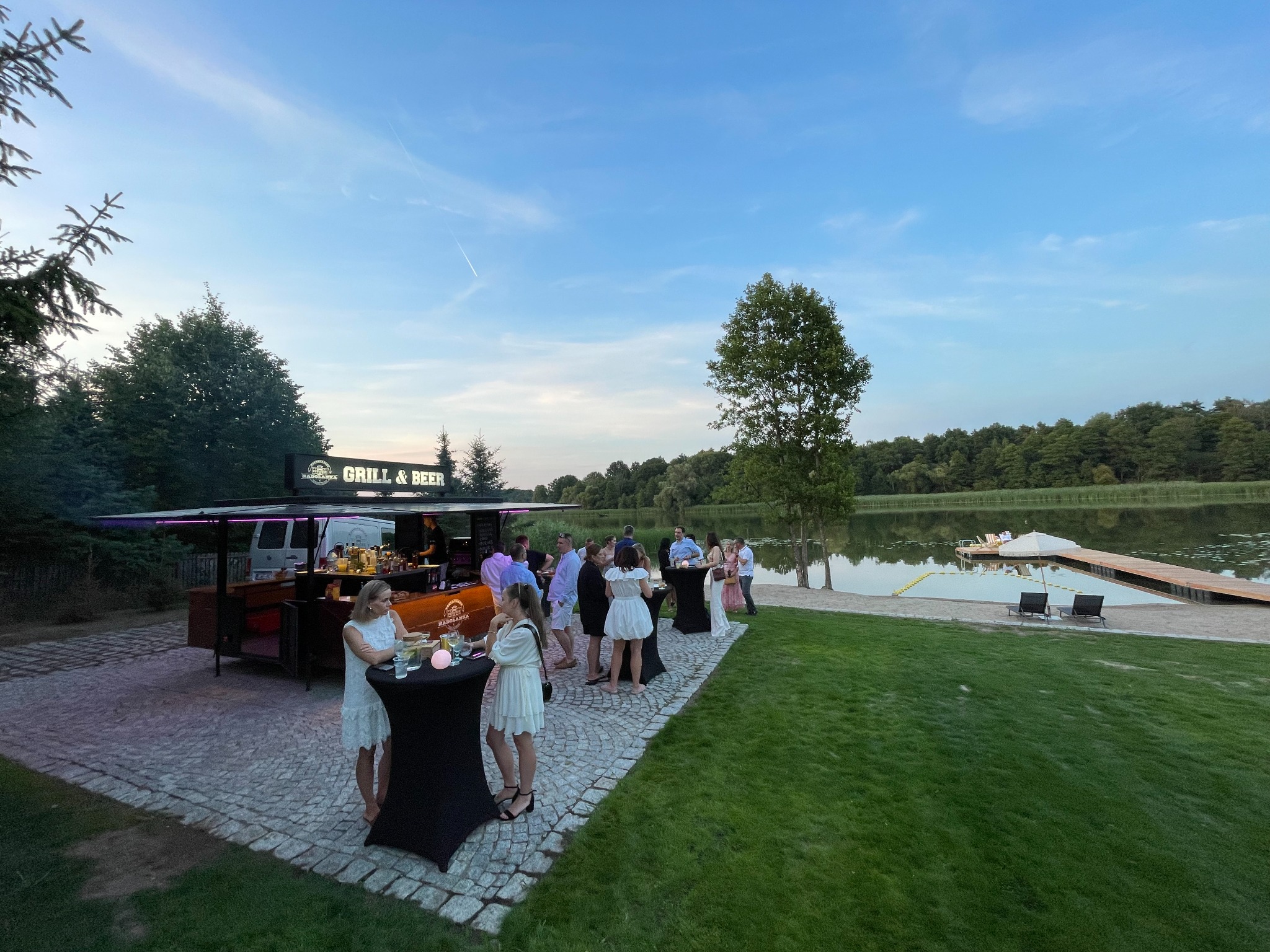 Plenerowa impreza firmowa nad jeziorem: mobilny bar Grill & Beer, goście w strojach koktajlowych, stoliki z czarnymi obrusami, relaksacyjny klimat.