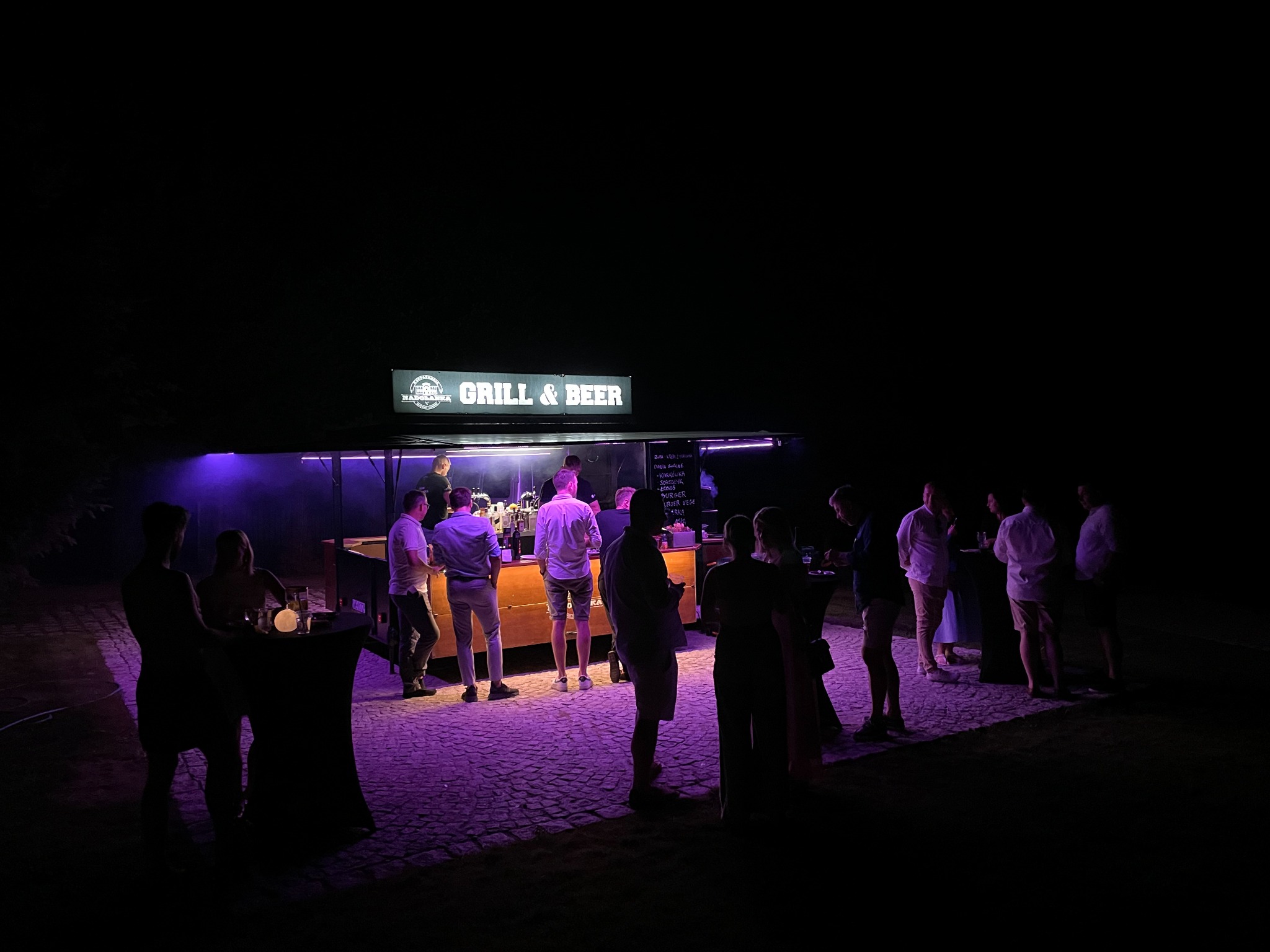 Mobilny bar Grill & Beer oświetlony fioletowym światłem, obsługa gości podczas wydarzenia na zewnątrz, widoczny bruk i sylwetki osób.