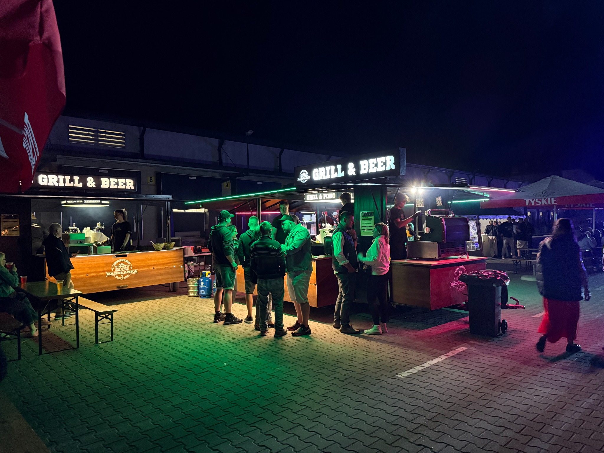 Nocne ujęcie stoisk 'Grill & Beer' z klientami, oświetlonych kolorowymi światłami, tworzącymi żywą atmosferę imprezy plenerowej. Widoczny fragment parasola Tyskie.