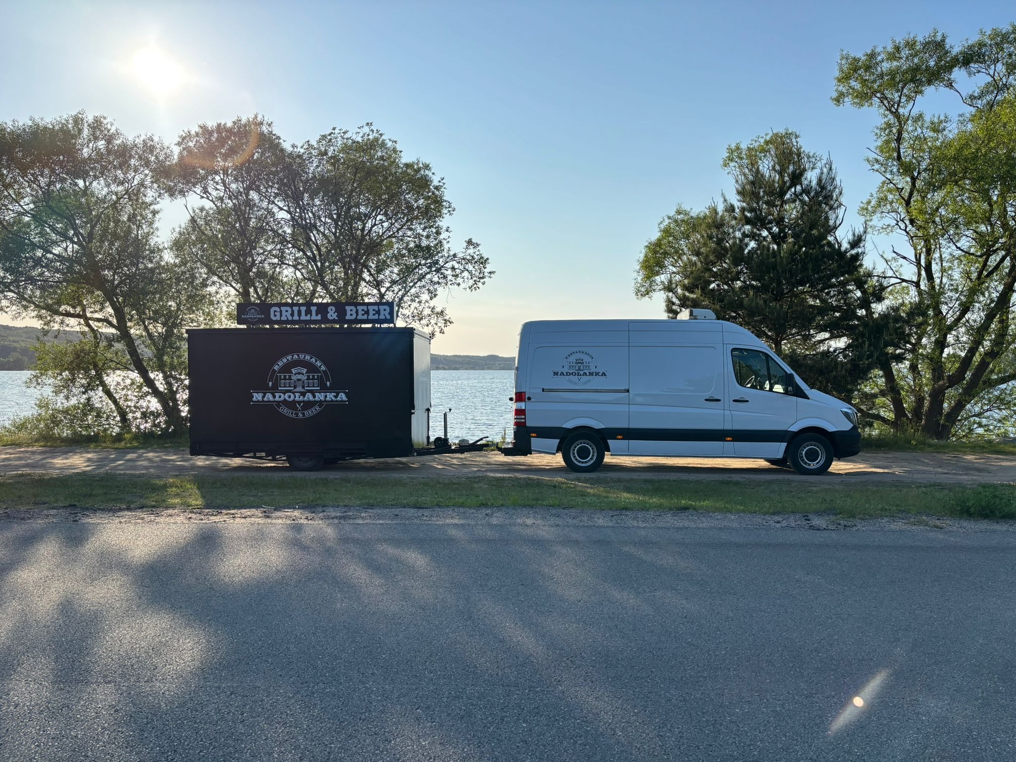 Mobilny catering 'Nadolanka Grill & Beer' z przyczepą na tle jeziora i zieleni. Biały van z logo firmy zaparkowany przy drodze.
