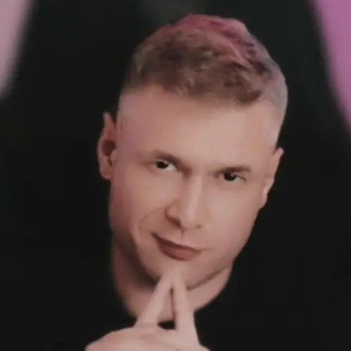 Portret mężczyzny z krótkimi włosami, w czarnej koszuli, z rękami złożonymi pod brodą, patrzącego wprost, na ciemnym tle z różowym akcentem.