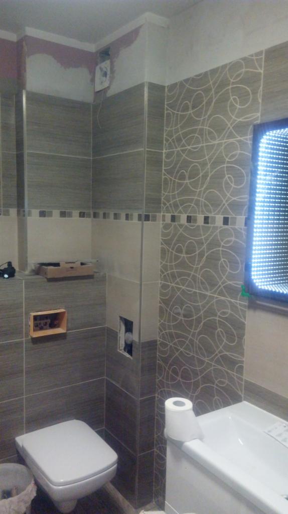 Wnętrze łazienki w trakcie wykończenia, z widoczną toaletą, wanną i częściowo ułożonymi płytkami w odcieniach szarości z wzorem. Widoczne przygotowania do montażu.