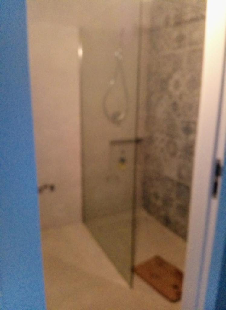 Szklana kabina prysznicowa z deszczownicą i brązową wycieraczką na podłodze, widok zza niebieskiej framugi drzwi.