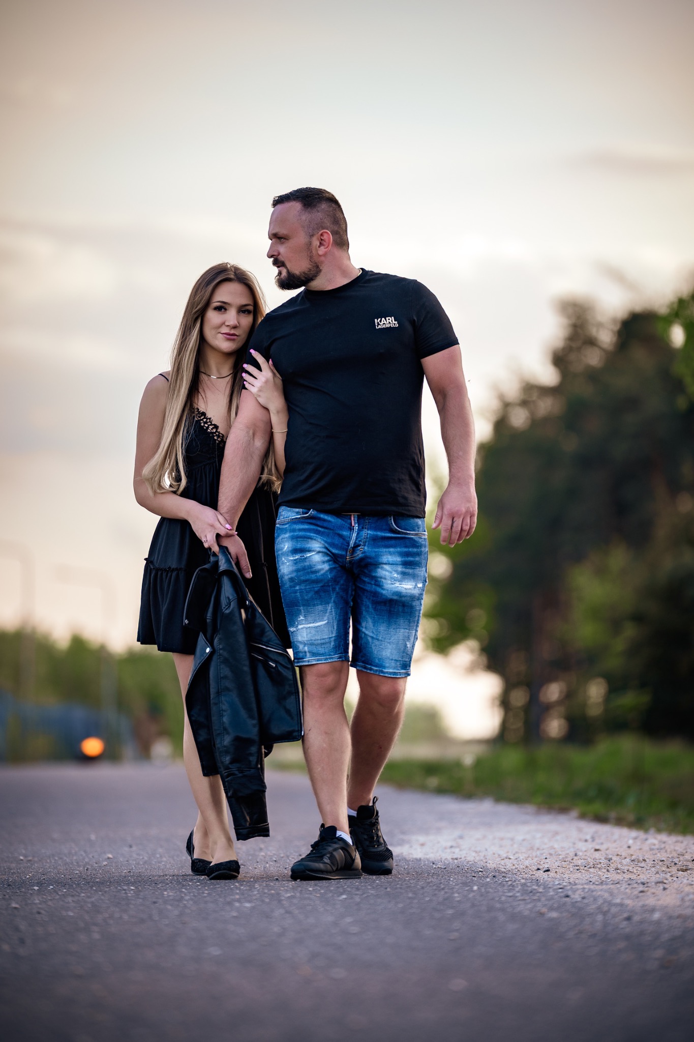 Zakochana para spaceruje po drodze o zmierzchu. Kobieta w czarnej sukience trzyma skórzaną kurtkę, mężczyzna w czarnym t-shircie i jeansowych szortach. Romantyczna sesja.