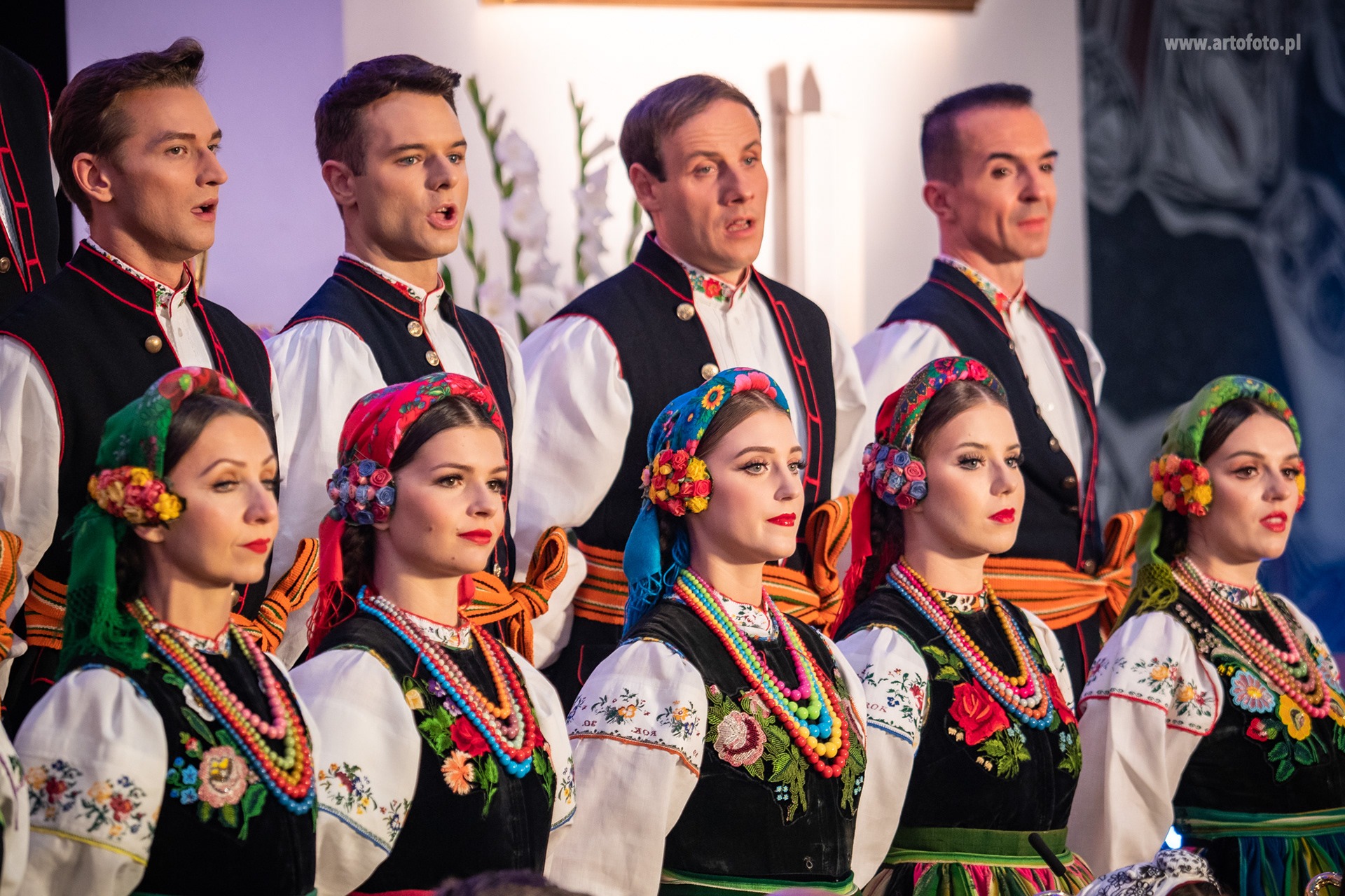 Zespół ludowy w tradycyjnych strojach śpiewa na scenie. Widoczne kolorowe, haftowane detale i kwiatowe nakrycia głowy. Jasne oświetlenie sceniczne.