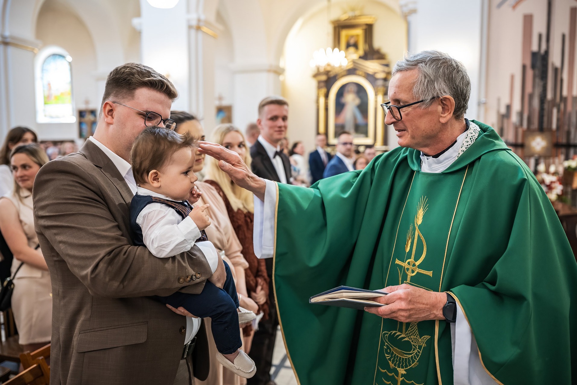 Ksiądz w zielonej szacie błogosławi małe dziecko trzymane przez ojca w kościele, w tle widoczni goście. Uroczystość religijna, fotografia z chrztu.