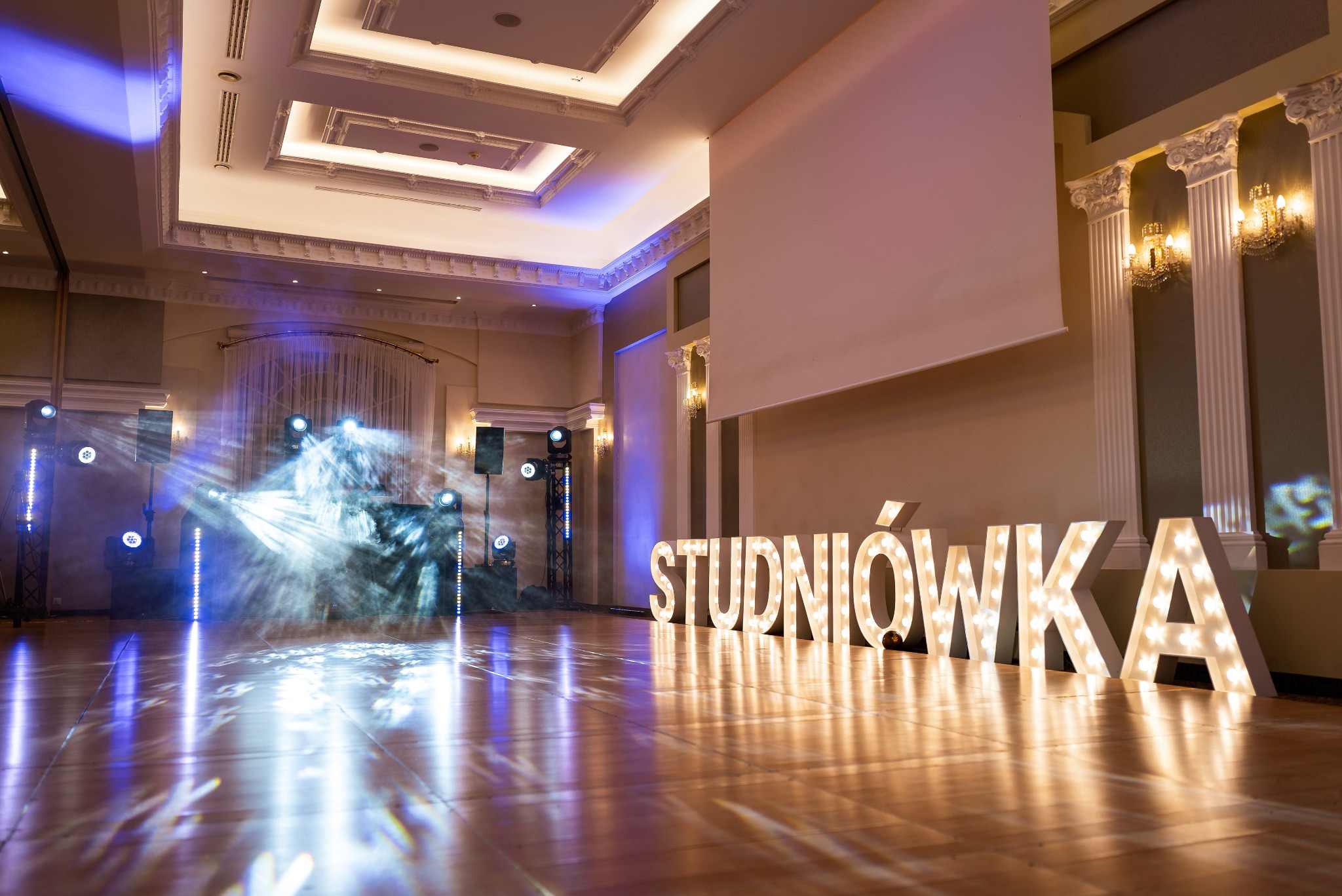 Sala balowa z dekoracją świetlną i dużym napisem 'STUDNIÓWKA' podświetlonym żarówkami. Parkiet odbija światła reflektorów.