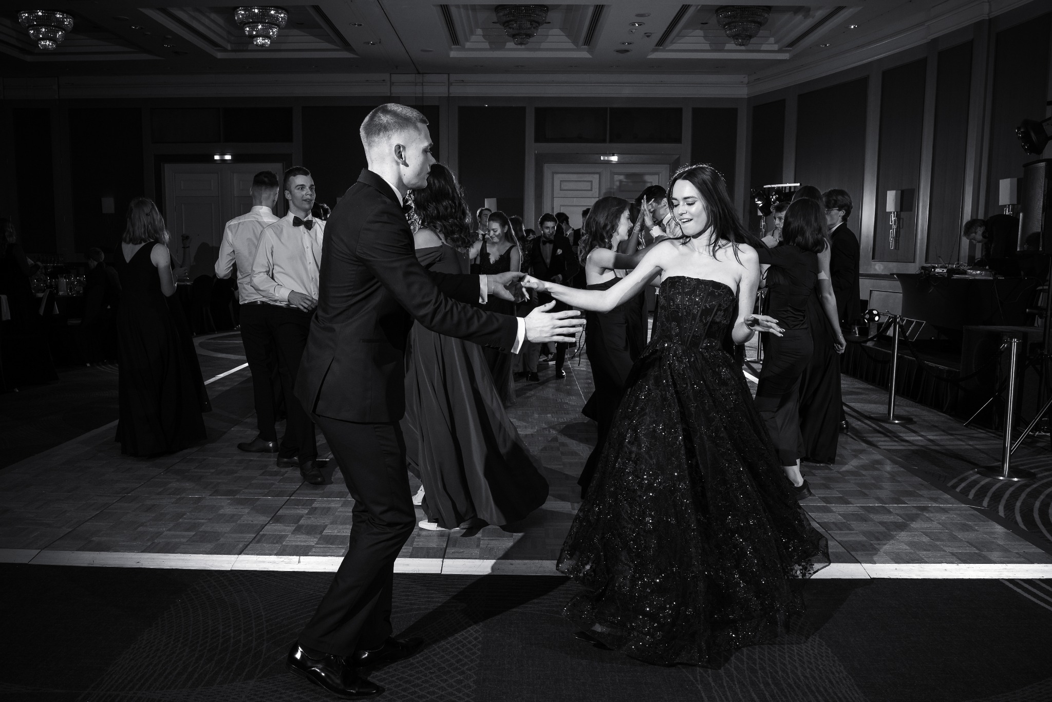Czarno-białe zdjęcie z balu. Para tańczy na parkiecie, wokół nich inni goście. Kobieta w błyszczącej sukni bez ramiączek, mężczyzna w garniturze. Uśmiechnięte twarze, radosna atmosfera.