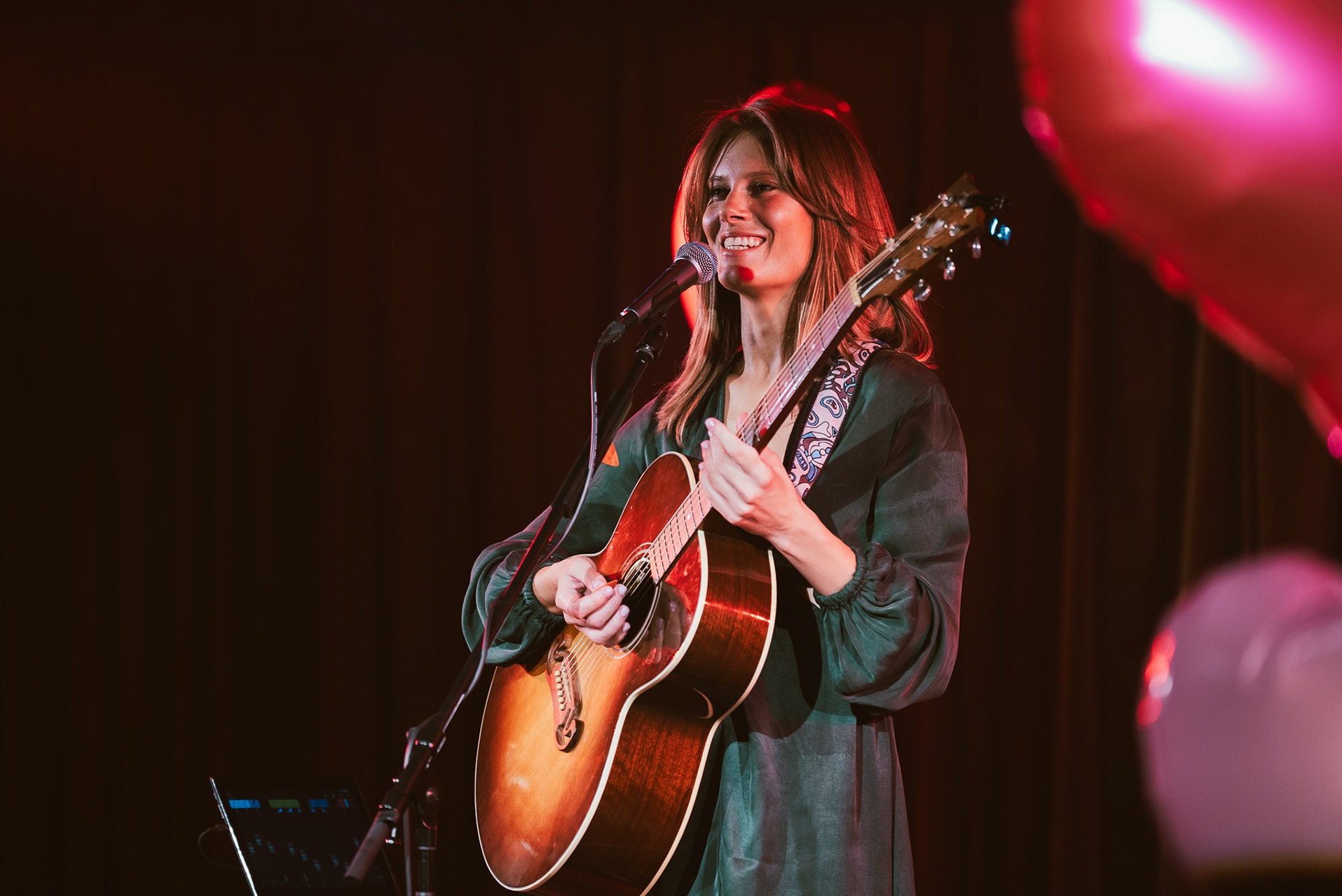 Uśmiechnięta kobieta w zielonej sukience śpiewa do mikrofonu, grając na akustycznej gitarze na scenie z ciemnym tłem i czerwonym oświetleniem.