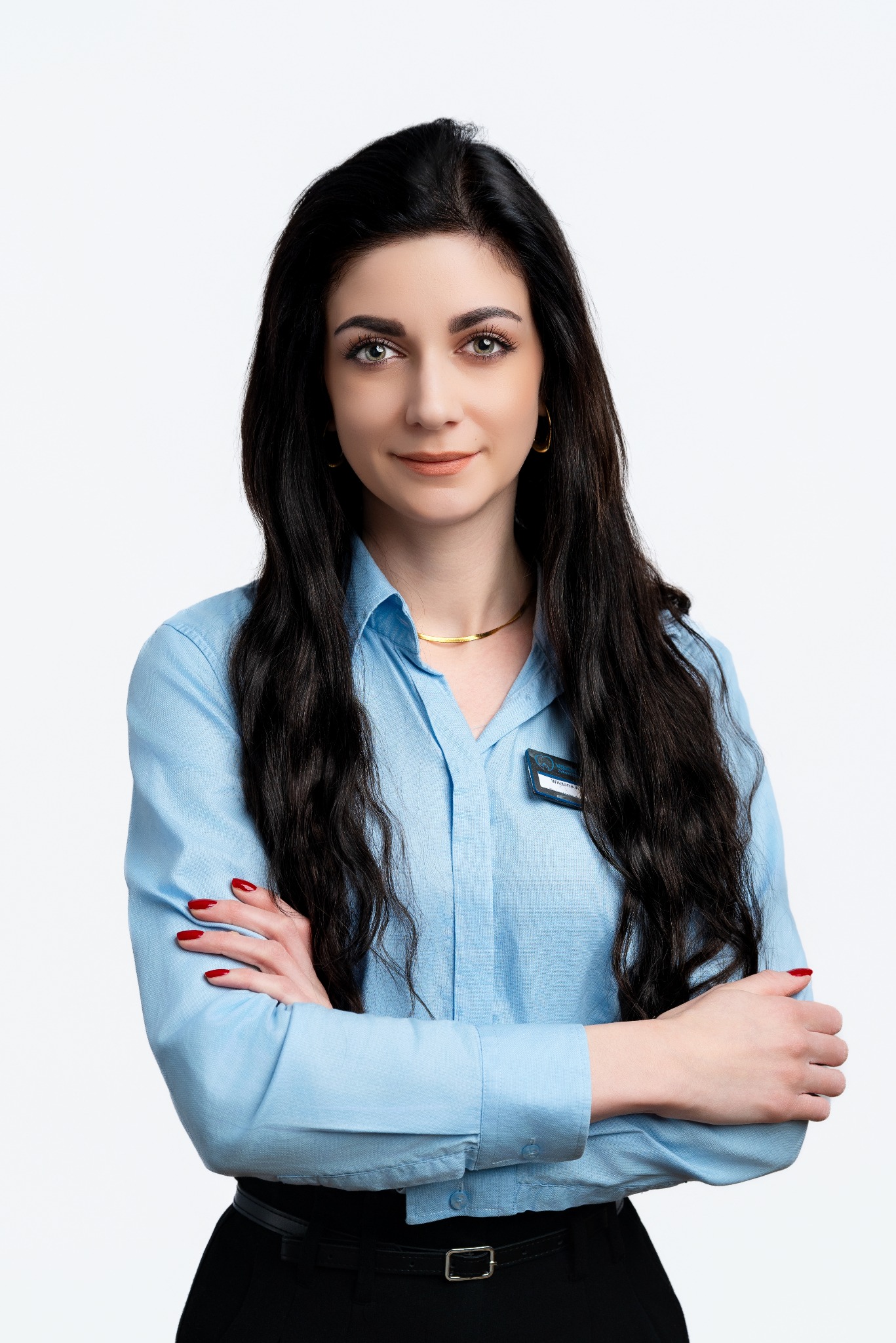 Portret kobiety z ciemnymi włosami, ubranej w błękitną koszulę, z założonymi rękami i identyfikatorem. Profesjonalny wizerunek na jasnym tle.