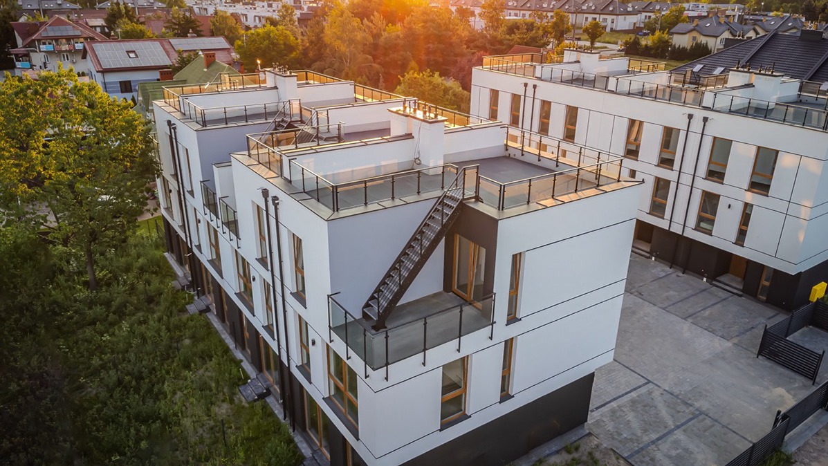 Nowoczesny budynek wielorodzinny z białymi elewacjami i szklanymi balustradami, widziany z góry w promieniach zachodzącego słońca. Schody na dachu.