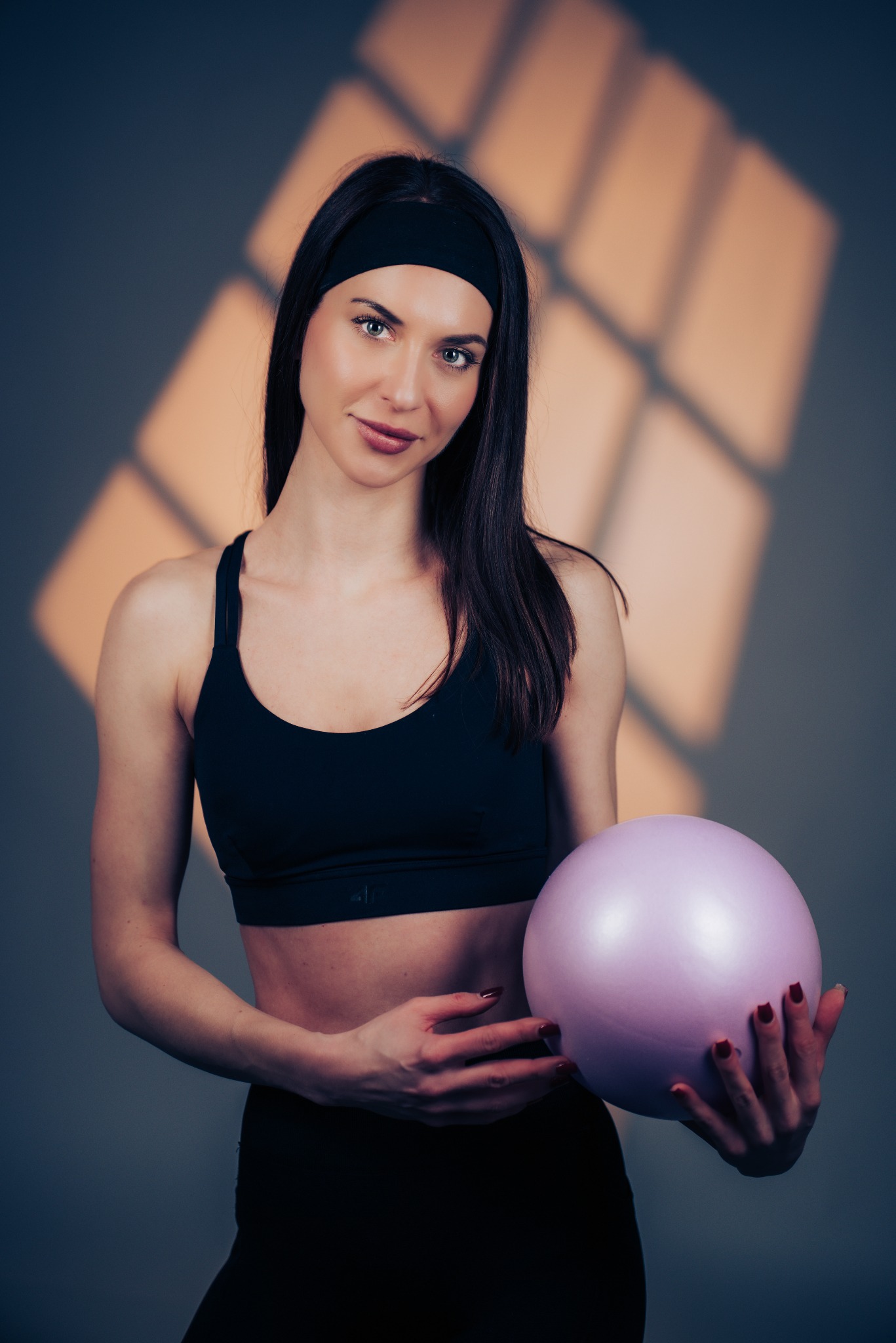Kobieta w sportowym stroju trzymająca fioletową piłkę do ćwiczeń na tle ściany z geometrycznym cieniem. Uśmiechnięta, z opaską na włosach, w czarnym topie i legginsach.