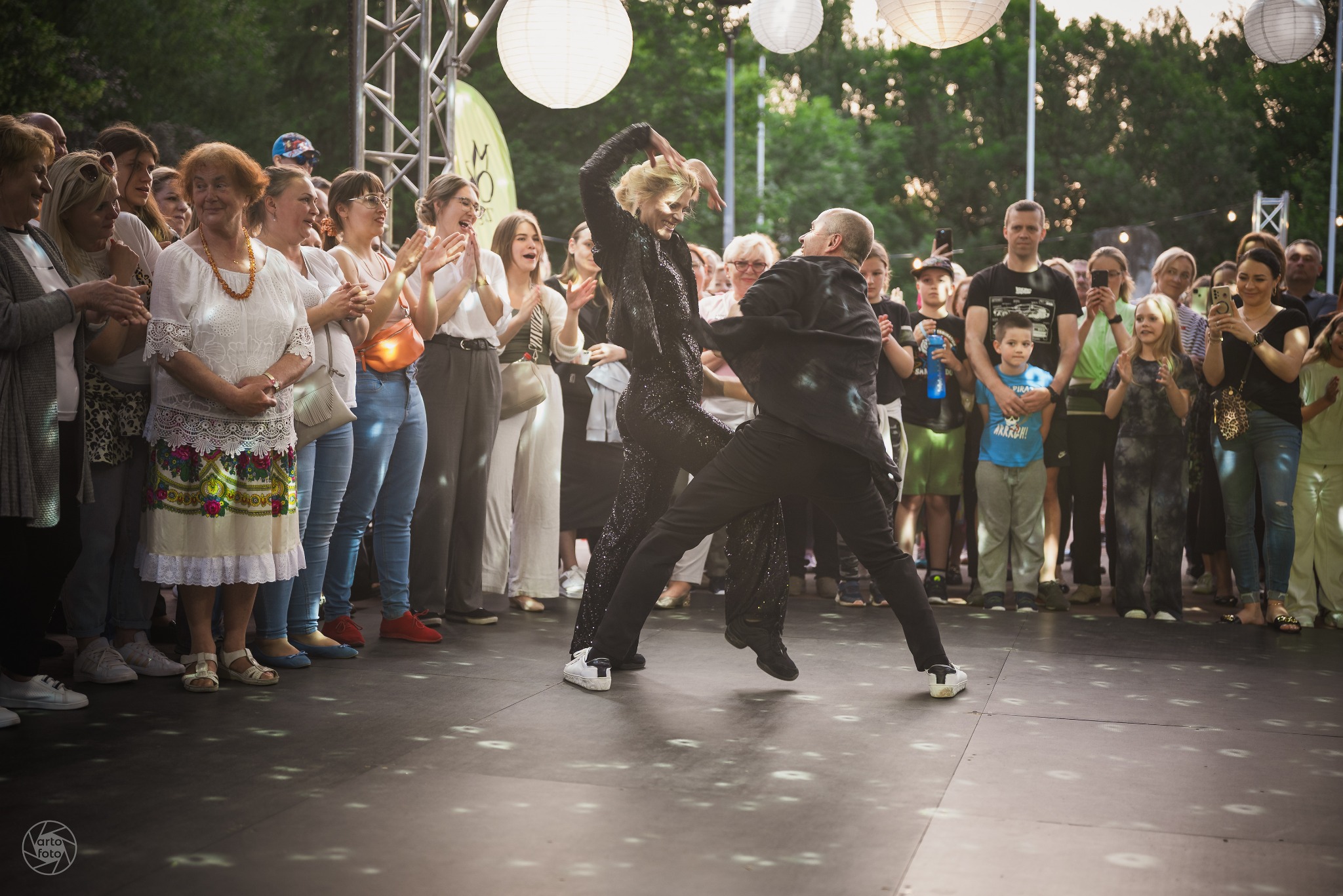 Para w dynamicznym tańcu na scenie, otoczona publicznością. Kobieta w cekinowym kombinezonie, mężczyzna w czarnym stroju. Scena oświetlona lampionami, widzowie z telefonami.