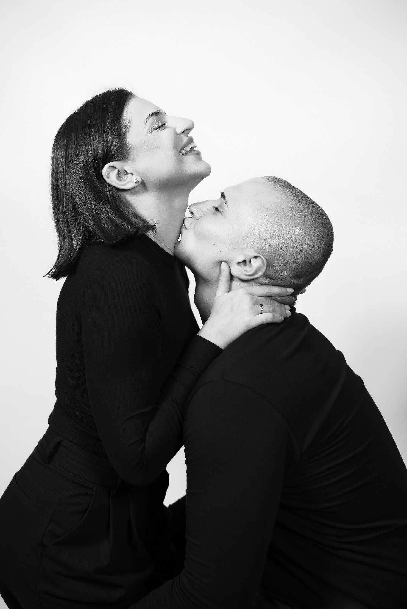 Czarno-białe zdjęcie pary: mężczyzna całuje kobietę w szyję, oboje ubrani na czarno, ona z uśmiechem, w tle jasne tło. Widoczny pierścionek na palcu kobiety.