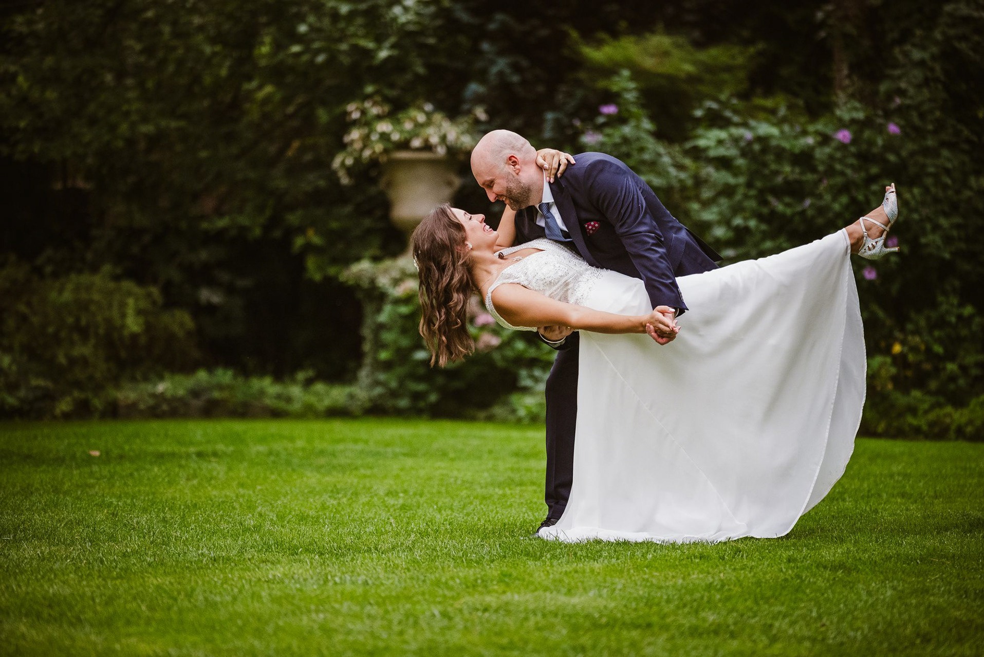 Para młoda w tańcu na tle zielonego ogrodu. Panna młoda w białej sukni i srebrnych butach, pan młody w granatowym garniturze. Uśmiechnięci, patrzą na siebie.