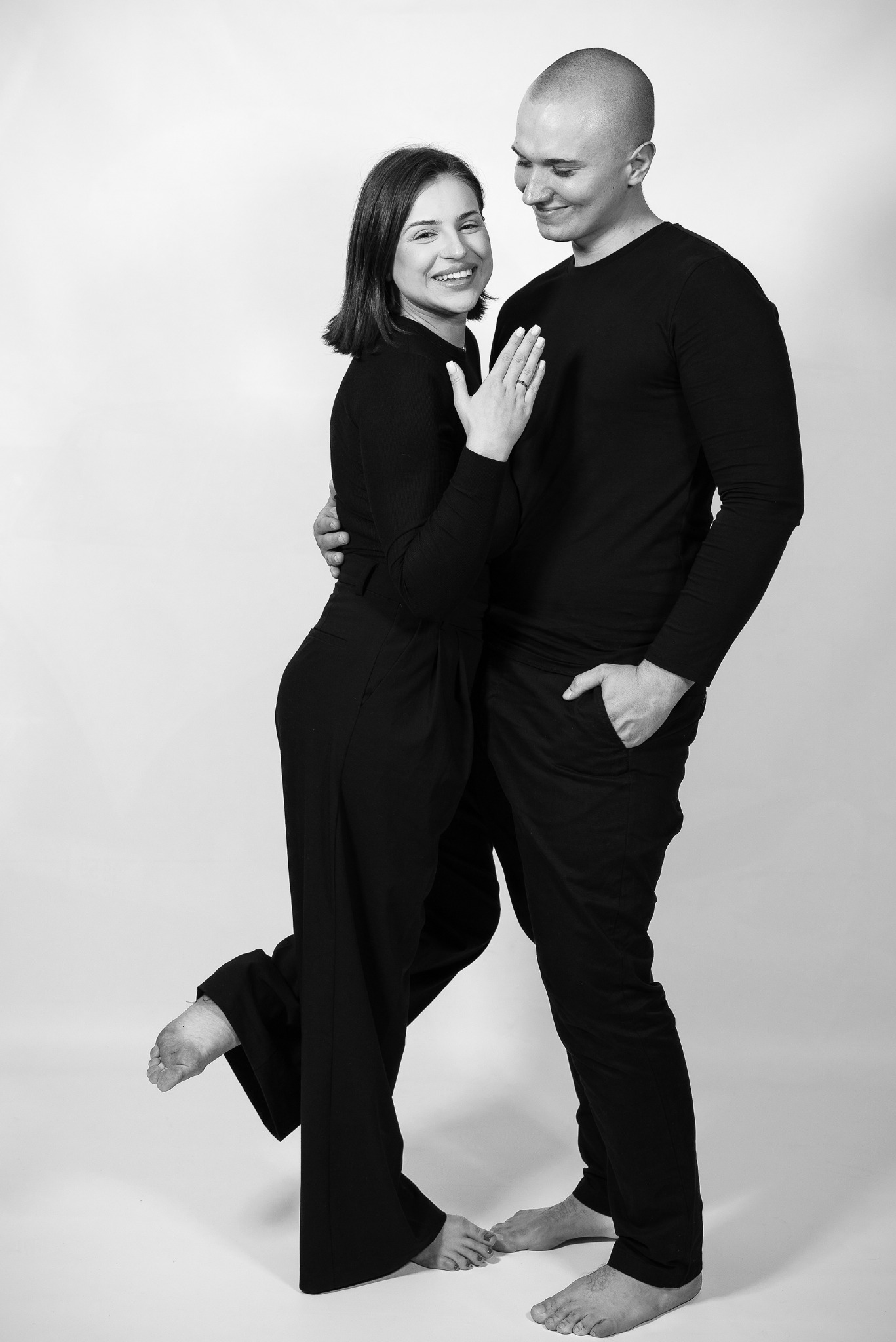 Czarno-białe, pełne ujęcie pary w czarnych strojach, kobieta z uniesioną nogą i pierścionkiem, mężczyzna z ręką w kieszeni, na jasnym tle. Sesja narzeczeńska.
