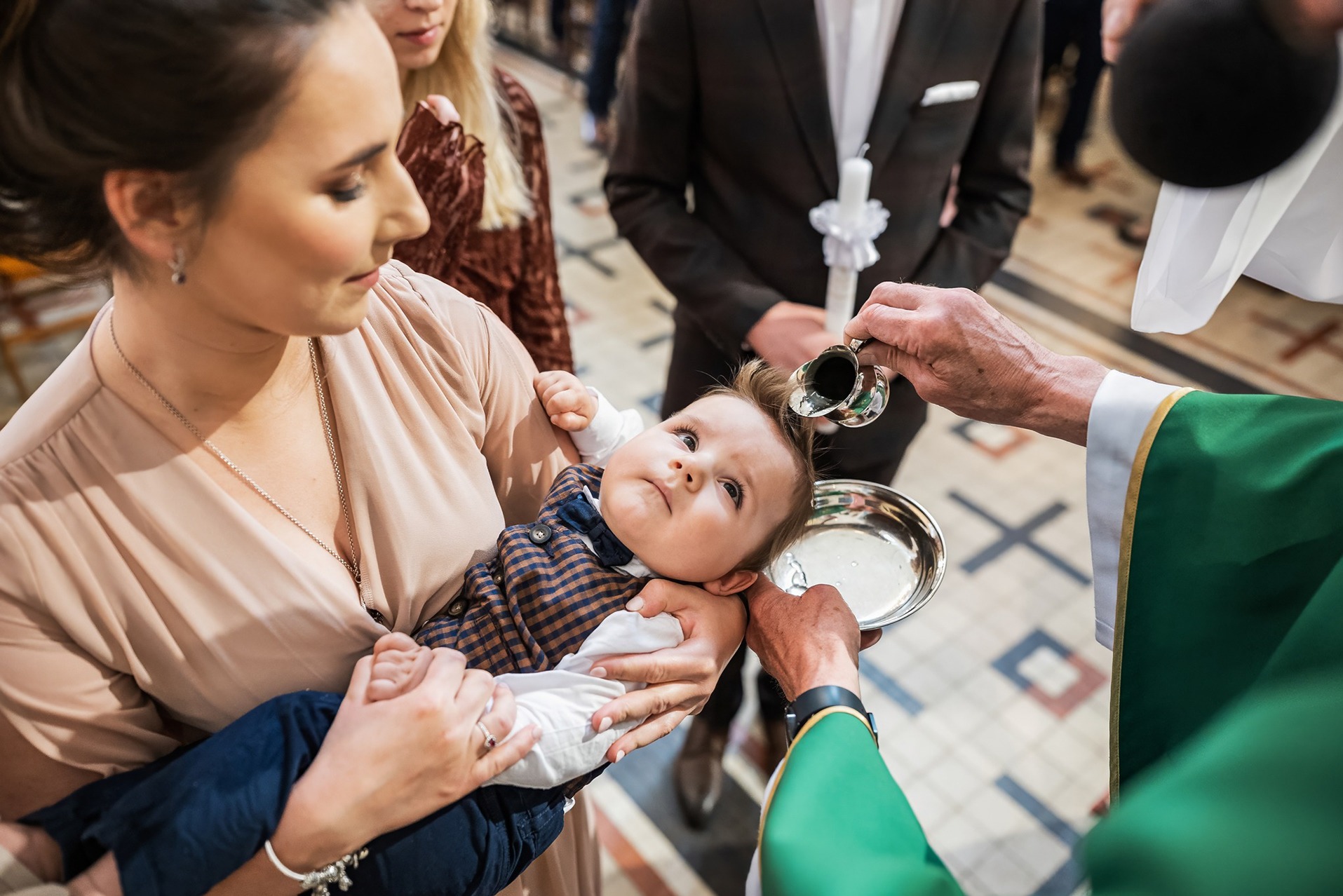 Chrzest dziecka: mama trzyma niemowlę w brązową kratę, ksiądz polewa wodą z naczynia. W tle goście, świeca. Ujęcie z bliska, naturalne światło.