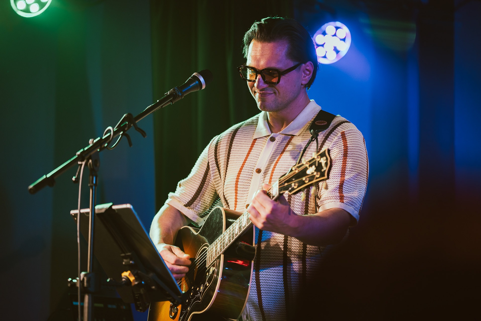 Mężczyzna w okularach gra na gitarze akustycznej na scenie, oświetlony zielonym i niebieskim światłem. Widoczny mikrofon i statyw na nuty. Uśmiechnięty muzyk w koszuli polo.