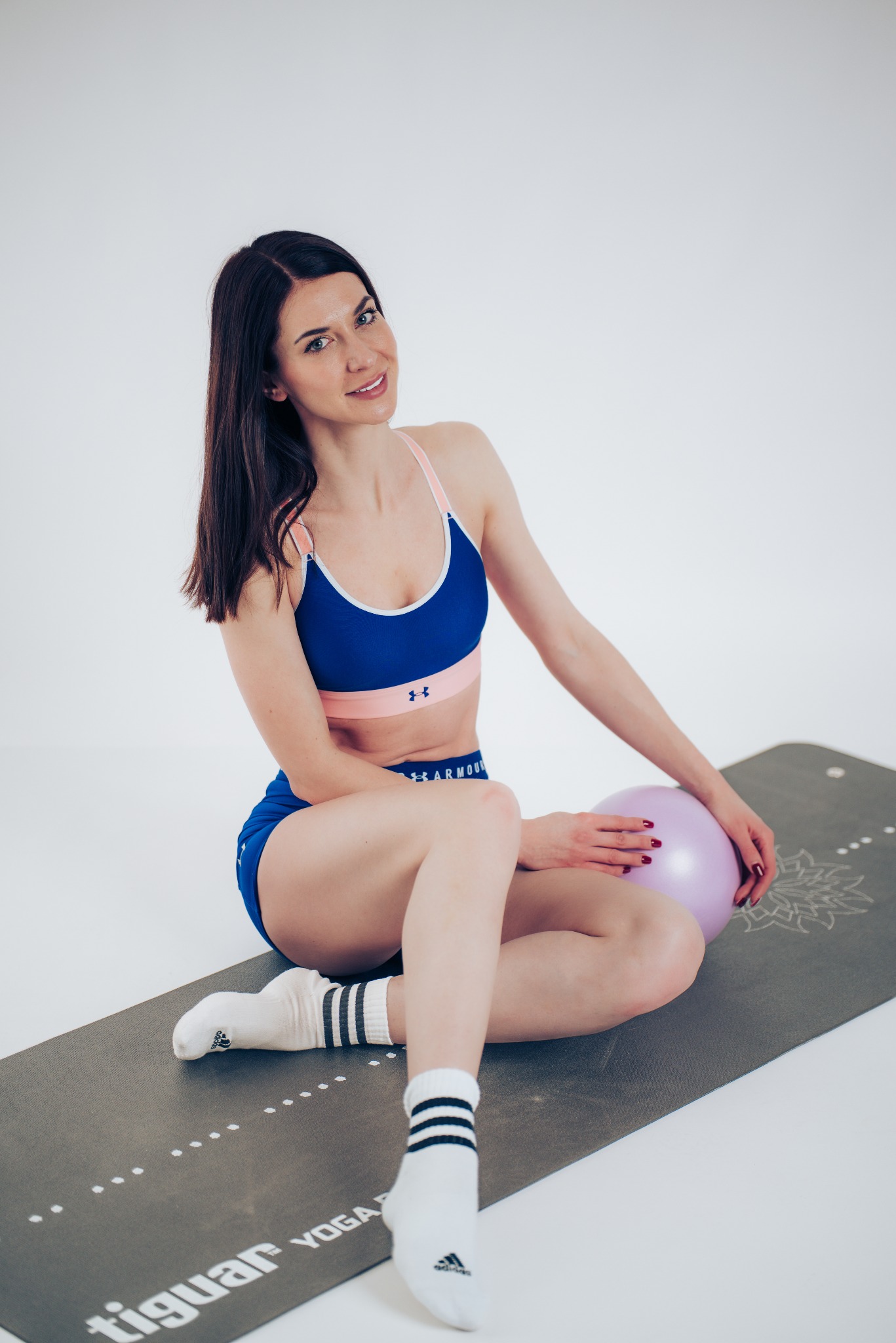 Kobieta w sportowym stroju siedzi na macie do jogi, trzymając fioletową piłkę. Uśmiechnięta brunetka w skarpetkach Adidas, na białym tle.