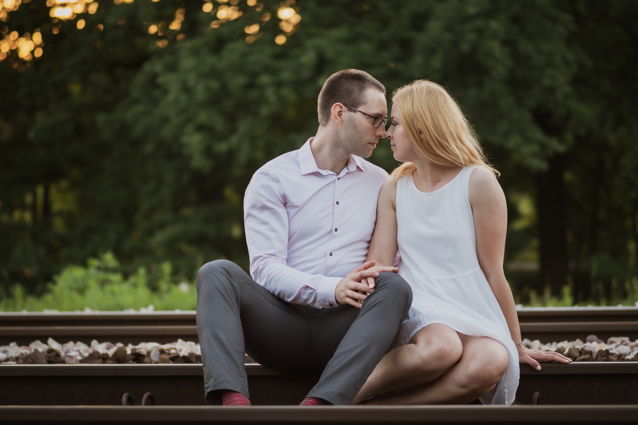 Zakochana para siedzi na torach kolejowych, blisko siebie, w tle zieleń drzew. Romantyczna sesja narzeczeńska w plenerze, delikatne światło.