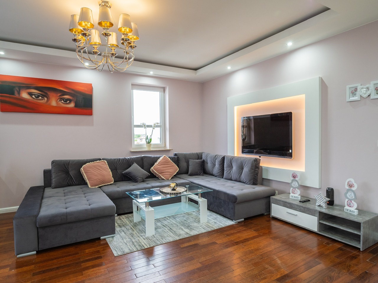 Nowoczesny salon z szarą sofą narożną, szklanym stolikiem kawowym i podświetlanym telewizorem. Na ścianie obraz z motywem oczu i dekoracyjna lampa sufitowa.