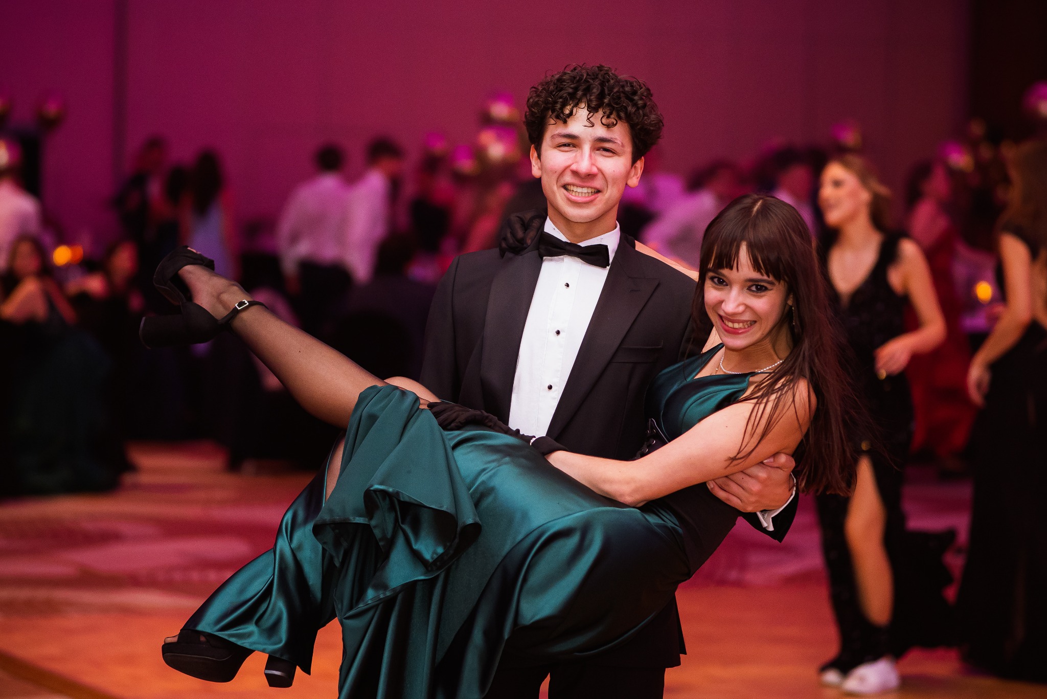 Uśmiechnięty mężczyzna w smokingu trzyma na rękach kobietę w długiej, zielonej sukni na tle rozmytego tłumu podczas uroczystości.