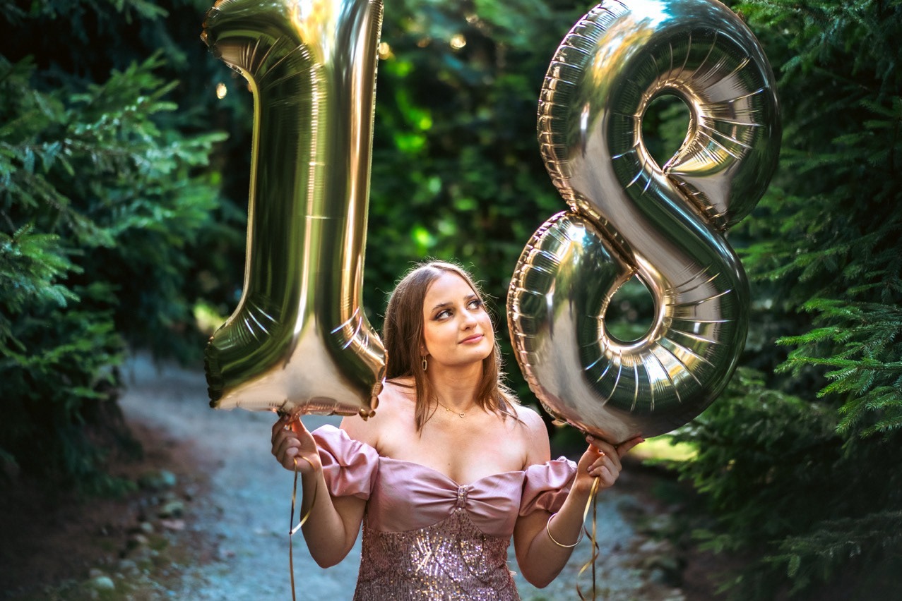 Uśmiechnięta osiemnastolatka w sukience trzyma złote balony w kształcie cyfr '1' i '8' na tle zieleni. Sesja urodzinowa w plenerze.