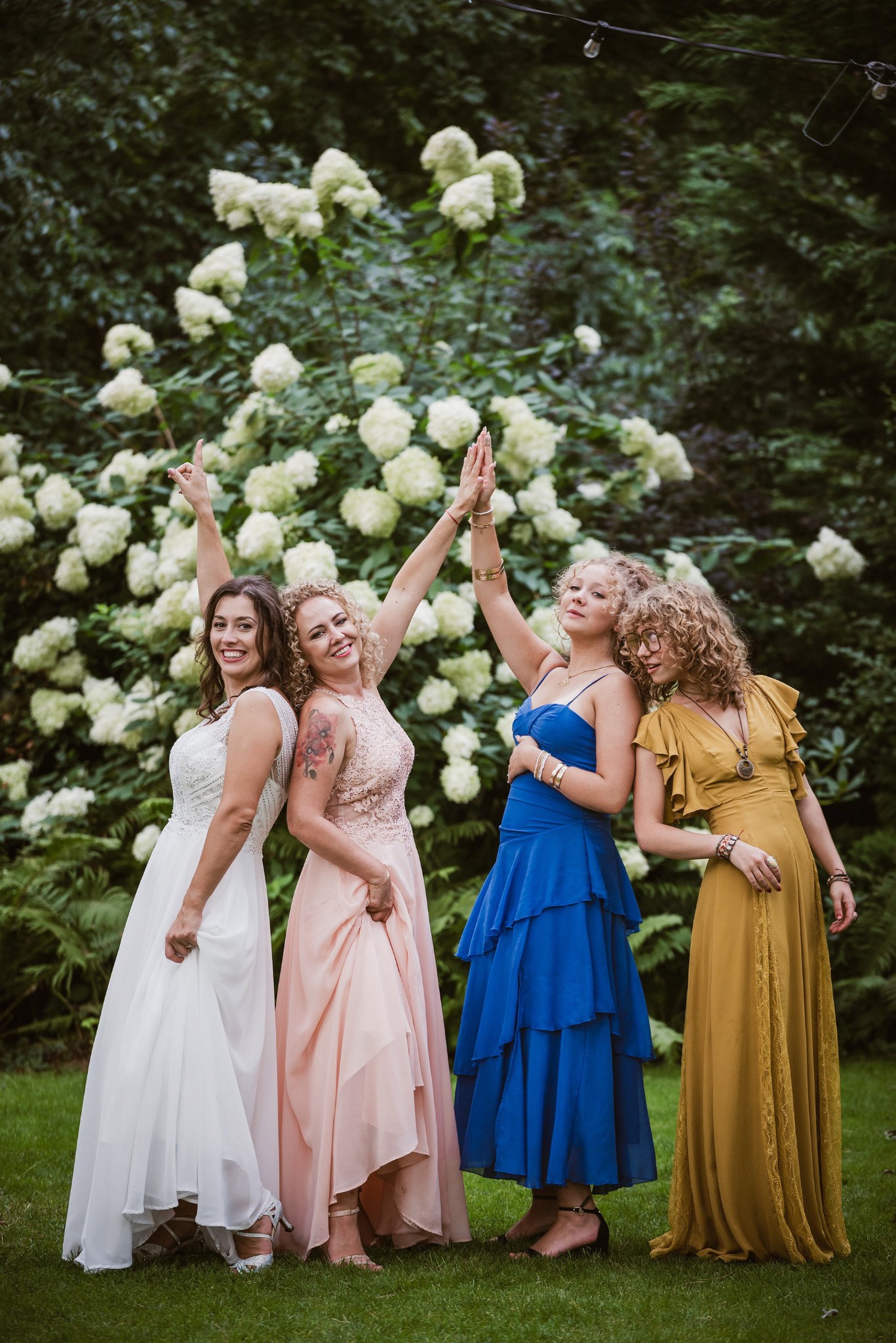 Cztery uśmiechnięte kobiety w sukniach wieczorowych, stojące tyłem do siebie na tle kwitnących hortensji, z podniesionymi rękami, w plenerze.