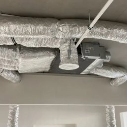 ZI System - Srebrne, elastyczne rury wentylacyjne zamontowane pod sufitem w jasnym pomieszczeniu. Widoczny centralny moduł wentylacyjny i fragment instalacji na ścianie.