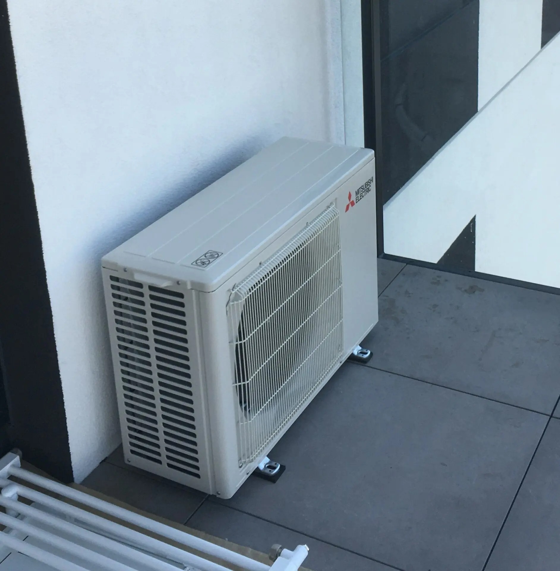 Biała jednostka zewnętrzna klimatyzacji Mitsubishi Electric na szarych płytkach balkonu, widoczna z góry pod kątem. Obok biała ściana i fragment czarno-białej konstrukcji.