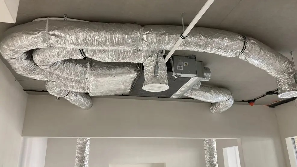 Srebrne, elastyczne rury wentylacyjne zamontowane pod sufitem w jasnym pomieszczeniu. Widoczny centralny moduł wentylacyjny i fragment instalacji na ścianie.