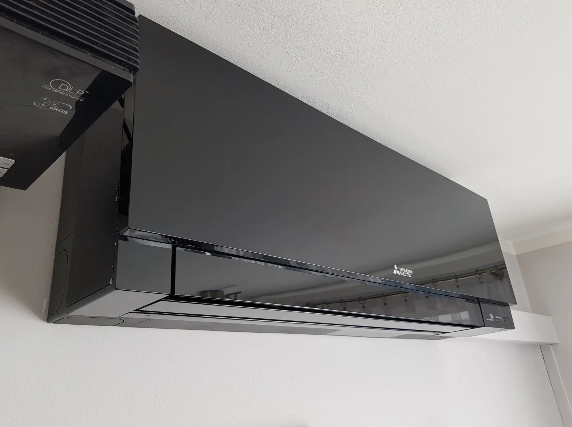 Czarny klimatyzator Mitsubishi Electric zamontowany na białej ścianie, widoczny z boku, z elementami instalacji elektrycznej w tle. Nowoczesny design.