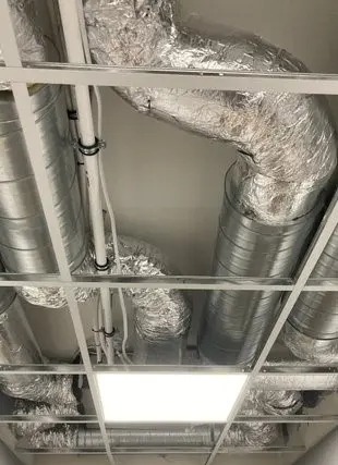 Srebrne, izolowane rury wentylacyjne podwieszone pod sufitem rastrowym z widocznym oświetleniem LED. Instalacja w budynku komercyjnym.