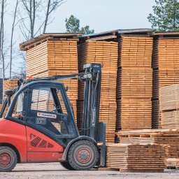 Neba, UAB - Czerwony wózek widłowy Linde na tle stosów jasnej tarcicy budowlanej. Widoczne drzewa w tle. Ujęcie z bliska, podkreślające strukturę drewna i detale wózka.