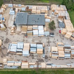 Neba, UAB - Widok z góry na skład drewna z budynkiem biurowym, stertami tarcicy budowlanej, drewnem kominkowym i wózkami widłowymi. Część drewna zabezpieczona folią.