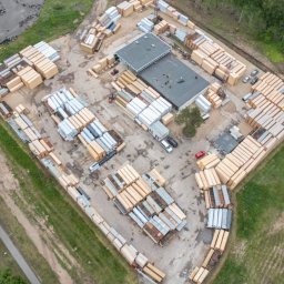 Neba, UAB - Widok z lotu ptaka na plac składowy z ułożonymi pakietami tarcicy budowlanej, z budynkiem biurowym w centrum i zielenią w tle. Widoczny wózek widłowy.