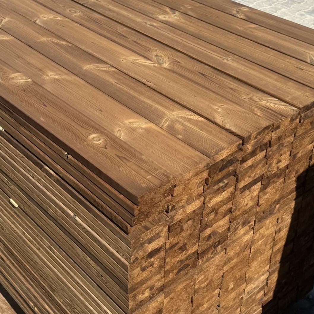 Stos desek tarasowych z termo sosny o gładkiej powierzchni, ułożonych warstwami. Widoczne słoje drewna i ciepły, brązowy odcień. Deski ułożone równo, tworząc stabilną konstrukcję.