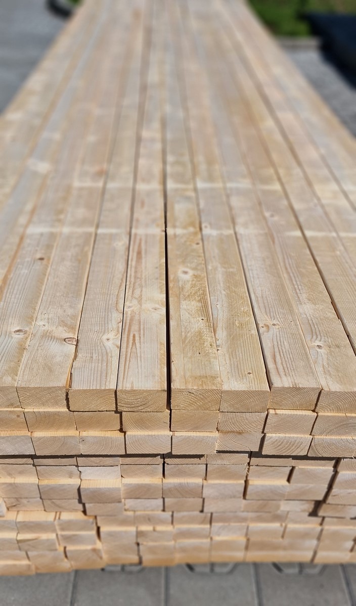 Stos belek z jasnego drewna C24 45x70, ułożonych równo jeden na drugim. Widoczne słoje drewna i krawędzie belek. Perspektywa z dołu, lekko zamazane tło.