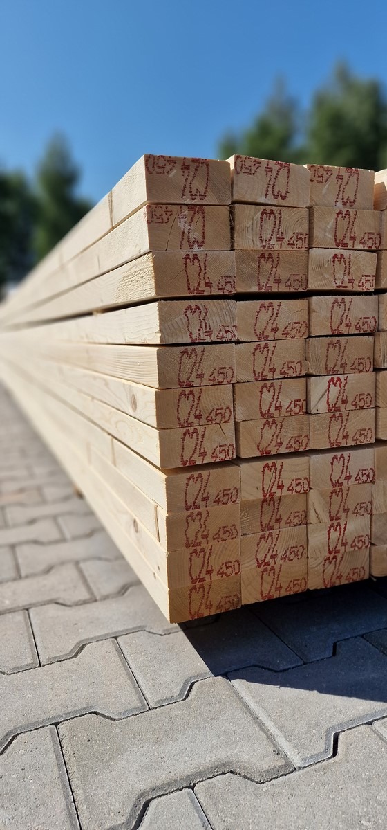 Stos tarcicy C24 45x95 ułożony na szarej kostce brukowej. Widoczne oznaczenia na końcach desek, błękitne niebo w tle. Jasne, naturalne oświetlenie.