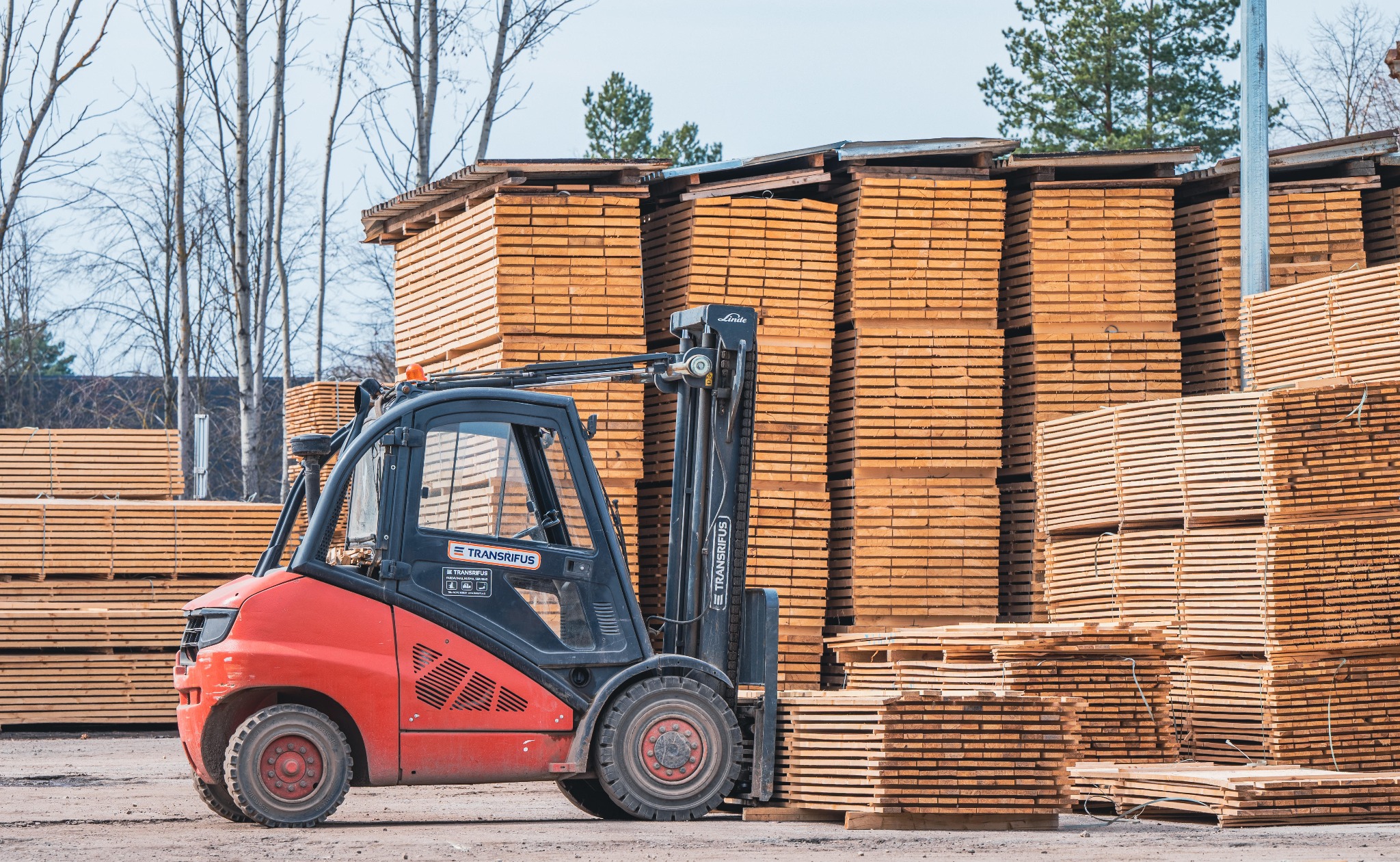 Czerwony wózek widłowy Linde na tle stosów jasnej tarcicy budowlanej. Widoczne drzewa w tle. Ujęcie z bliska, podkreślające strukturę drewna i detale wózka.