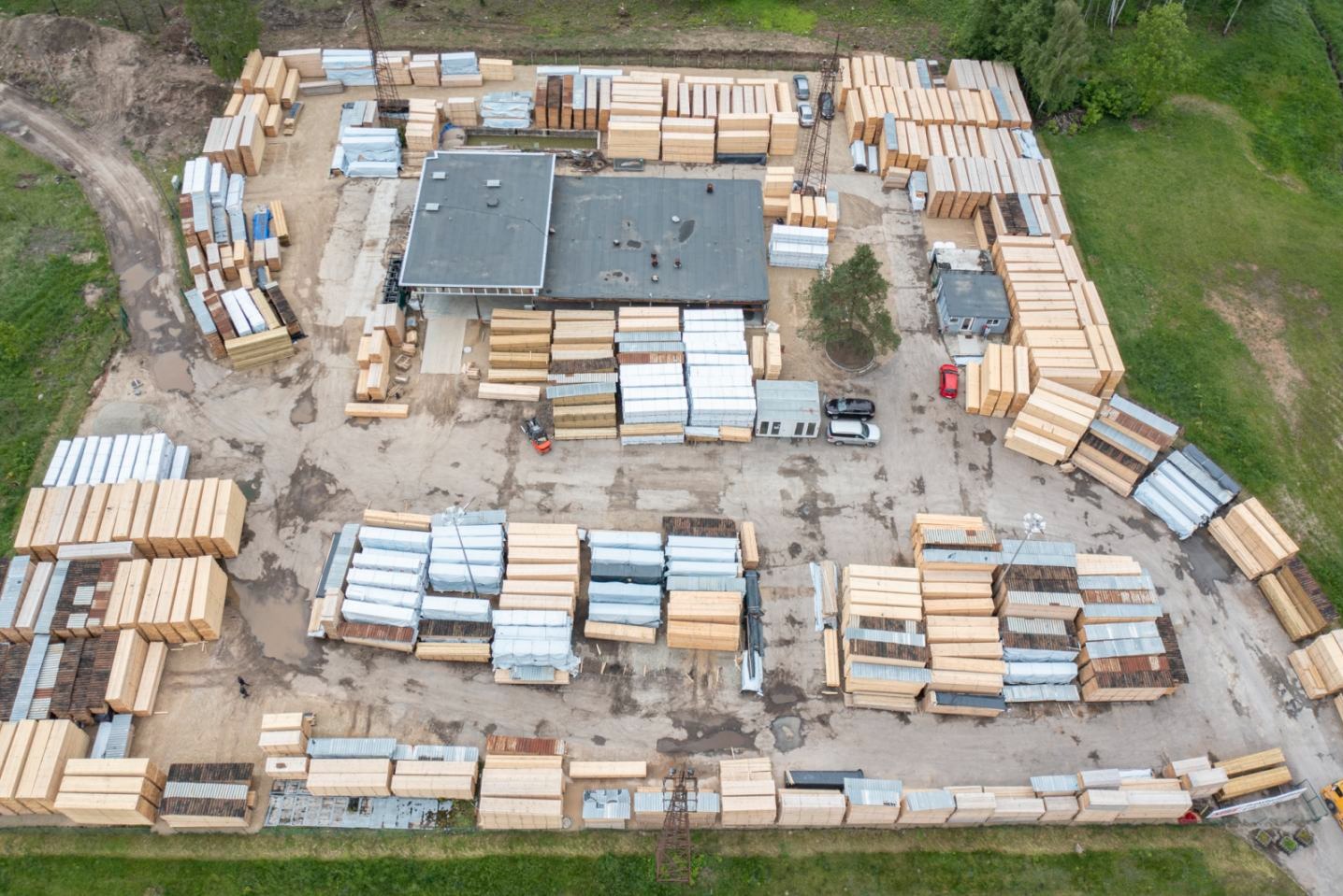 Widok z góry na skład drewna z budynkiem biurowym, stertami tarcicy budowlanej, drewnem kominkowym i wózkami widłowymi. Część drewna zabezpieczona folią.