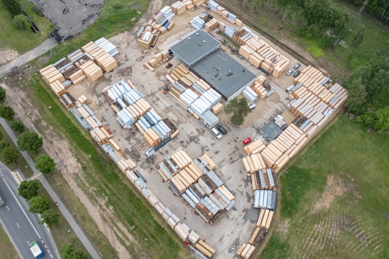 Widok z lotu ptaka na plac składowy z ułożonymi pakietami tarcicy budowlanej, z budynkiem biurowym w centrum i zielenią w tle. Widoczny wózek widłowy.