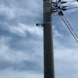 MeleM - Słup elektryczny z zamontowaną kamerą monitoringu na tle pochmurnego nieba. Widoczne przewody i elementy mocujące.