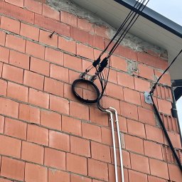 MeleM - Czarne kable elektryczne przymocowane do ściany z czerwonej cegły. Widoczne białe rurki PCV i fragment dachu. Ujęcie z dołu, pochmurne niebo w tle.
