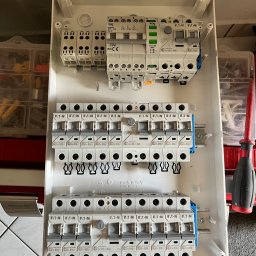 MeleM - Otwarta, biała skrzynka rozdzielcza z zamontowanymi bezpiecznikami EATON i IDEAL, widok z góry, ukazujący szczegóły instalacji elektrycznej.