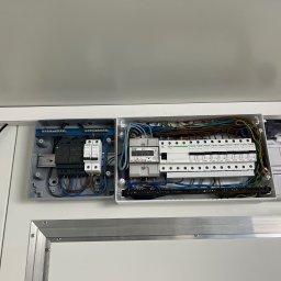 MeleM - Otwarta skrzynka elektryczna z bezpiecznikami, przewodami i licznikiem, zamontowana na białej ścianie. Widoczne elementy instalacji elektrycznej.